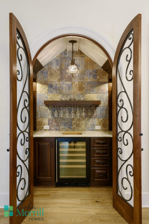 A stylish kitchen wet bar featuring a wine cooler, decorative tile backsplash, and wooden cabinetry, framed by elegant arched double doors with wrought iron details.