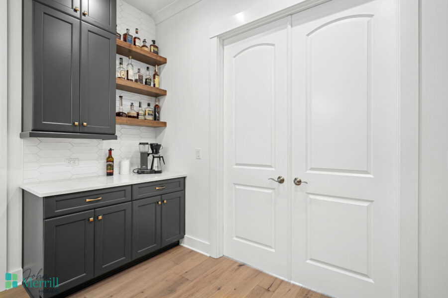 A stylish home bar area featuring dark gray cabinets, white countertops, and open wooden shelves displaying bottles and glassware.