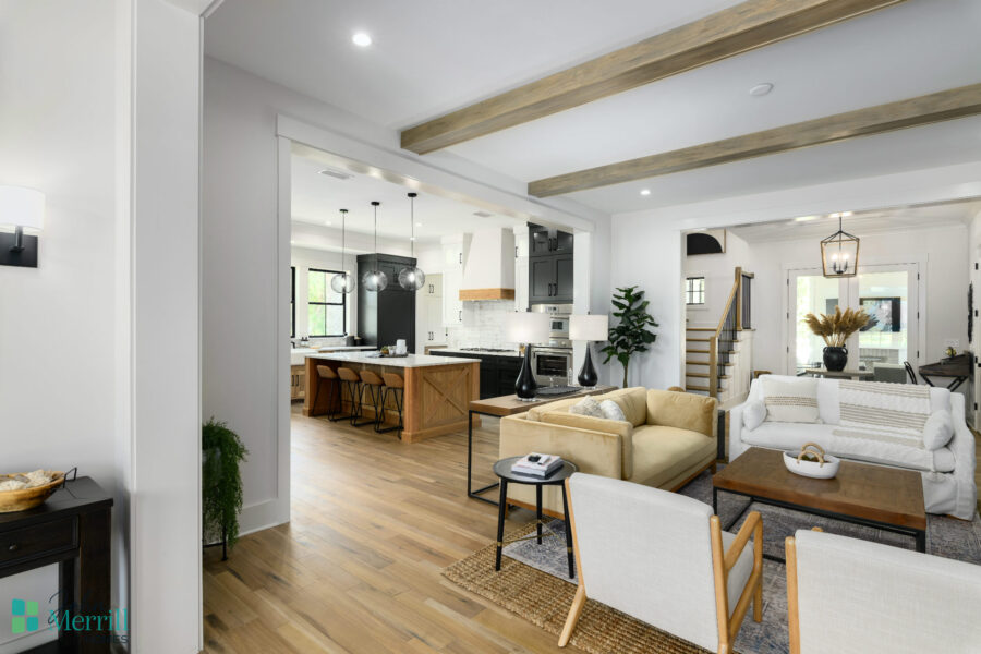 A spacious, bright living room with white walls, wooden beams, and large windows, seamlessly connected to a modern kitchen with an island and pendant lighting.