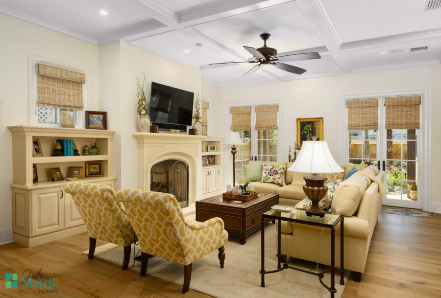 Bright and inviting living room with cream-colored sofas, yellow patterned armchairs, and a cozy fireplace, featuring large windows with woven shades and a ceiling fan.