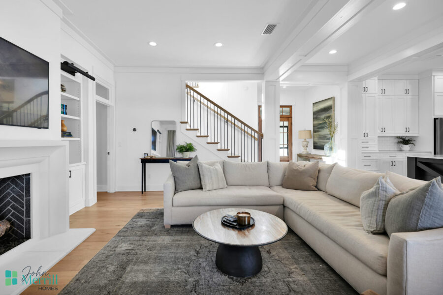 Bright and spacious living room featuring a large L-shaped sofa, a round coffee table, and a fireplace, with natural light streaming through windows and a staircase leading upstairs.