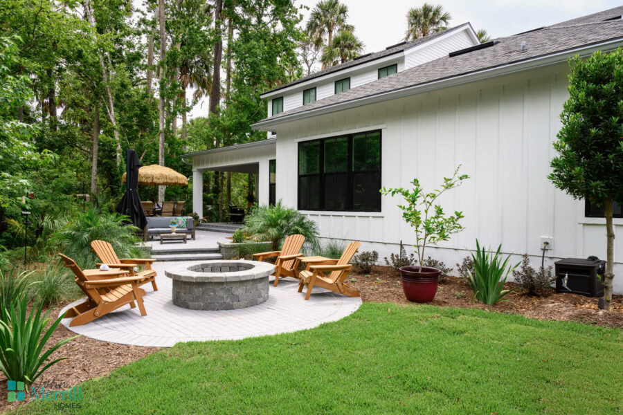A spacious backyard featuring a modern white house with large windows, a cozy outdoor seating area with Adirondack chairs around a fire pit, surrounded by lush greenery and tall trees.