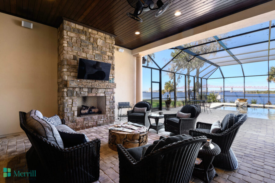 A covered patio featuring comfortable black wicker furniture, a stone fireplace, and a view of the water through a screened enclosure, perfect for relaxing and entertaining.