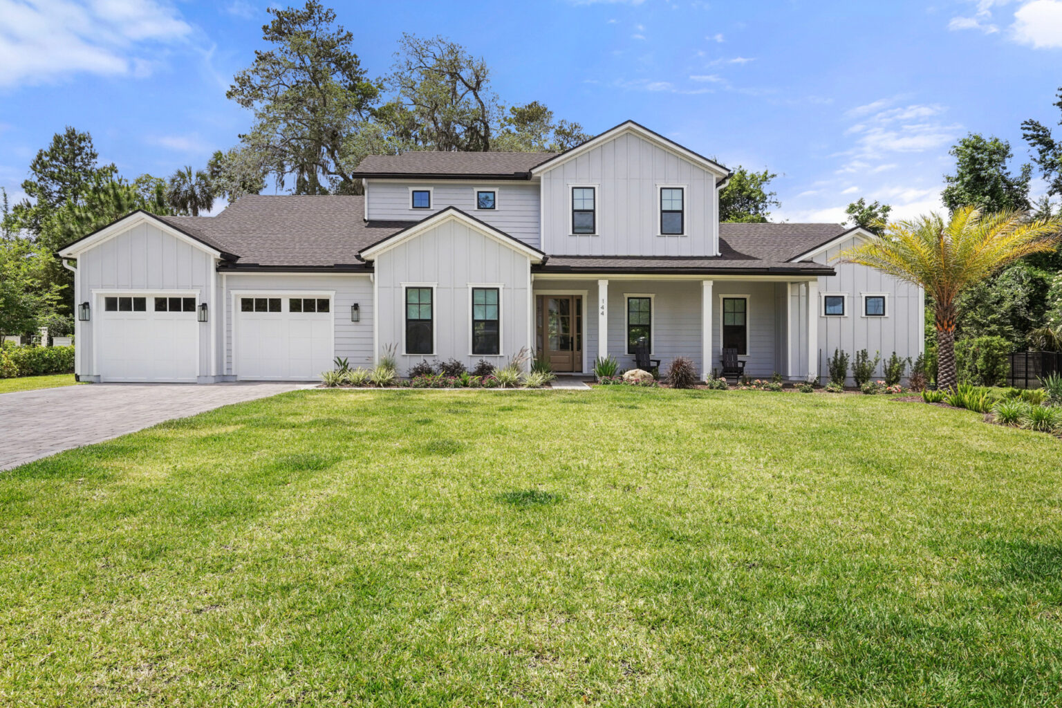 Luxurious modern white farmhouse-style home with a spacious front lawn, black window frames, and a three-car garage set against a bright blue sky and lush greenery.
