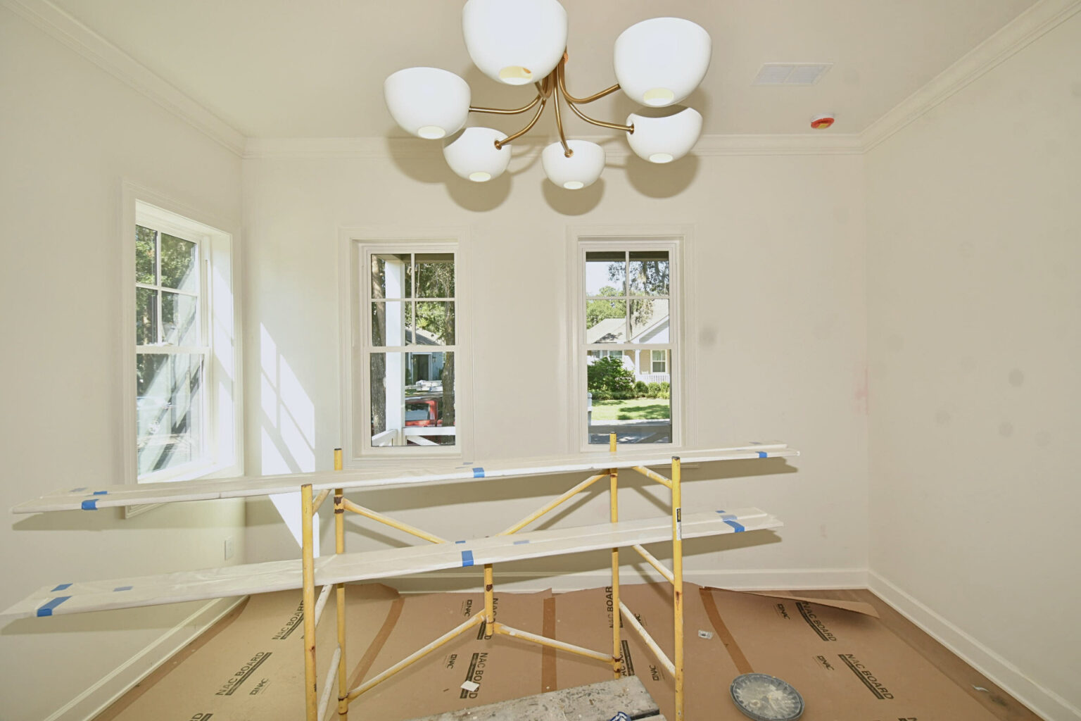 Brightly lit interior room with three large double-hung windows, white walls, a modern chandelier, and construction scaffolding. Perfect for showcasing a new home build by John Merrill Homes.