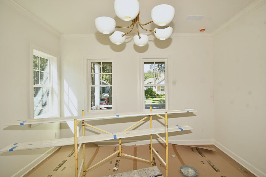 Brightly lit interior room with three large double-hung windows, white walls, a modern chandelier, and construction scaffolding. Perfect for showcasing a new home build by John Merrill Homes.