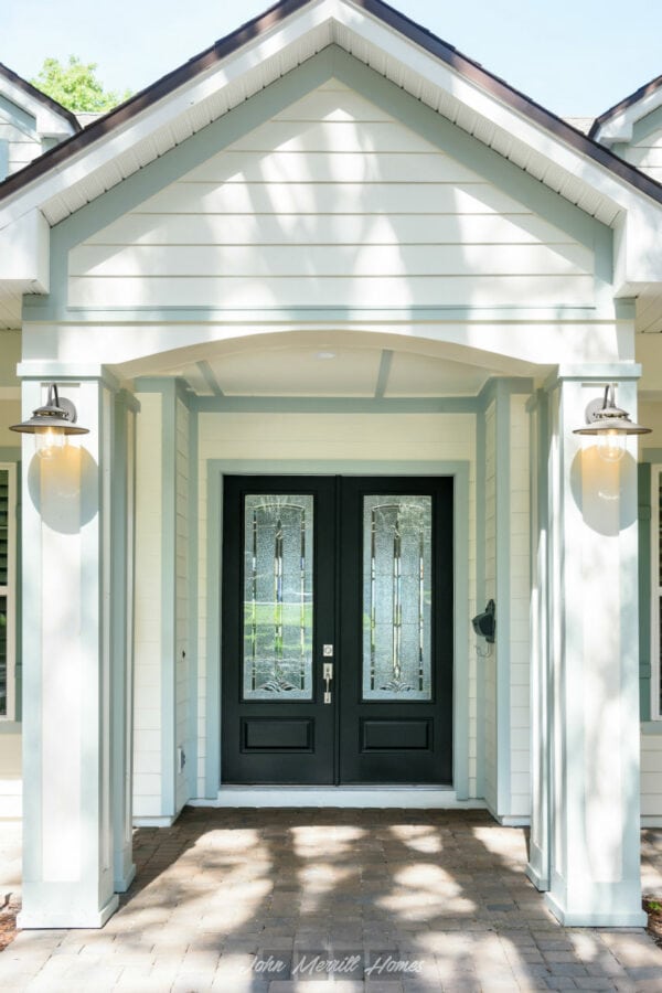 Black front door with decorative glass panels, white house exterior with porch lighting, and brick pathway, showcasing modern architectural design by John Merrill Homes.