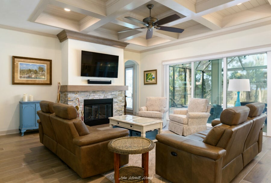 Cozy living room with leather sofas, a stone fireplace, a large flat-screen TV, and natural light streaming through sliding glass doors overlooking a lush outdoor area.