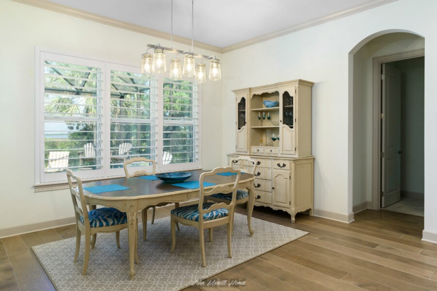 Bright dining room with large window shutters, cream-colored furniture, and a chandelier; opens to a scenic outdoor patio with greenery.