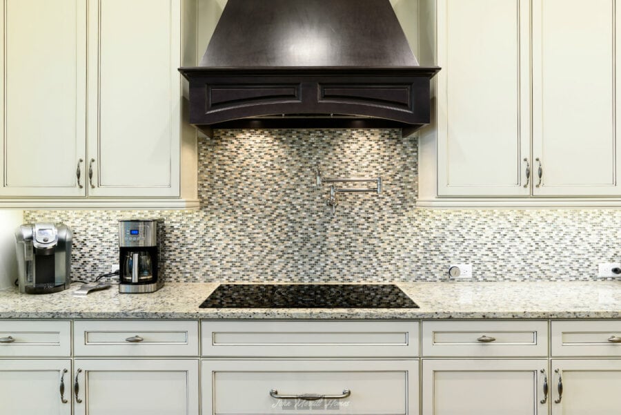 Polished kitchen with white cabinetry, granite countertops, modern appliances, and a mosaic tile backsplash above an electric cooktop with a stylish dark wood range hood.