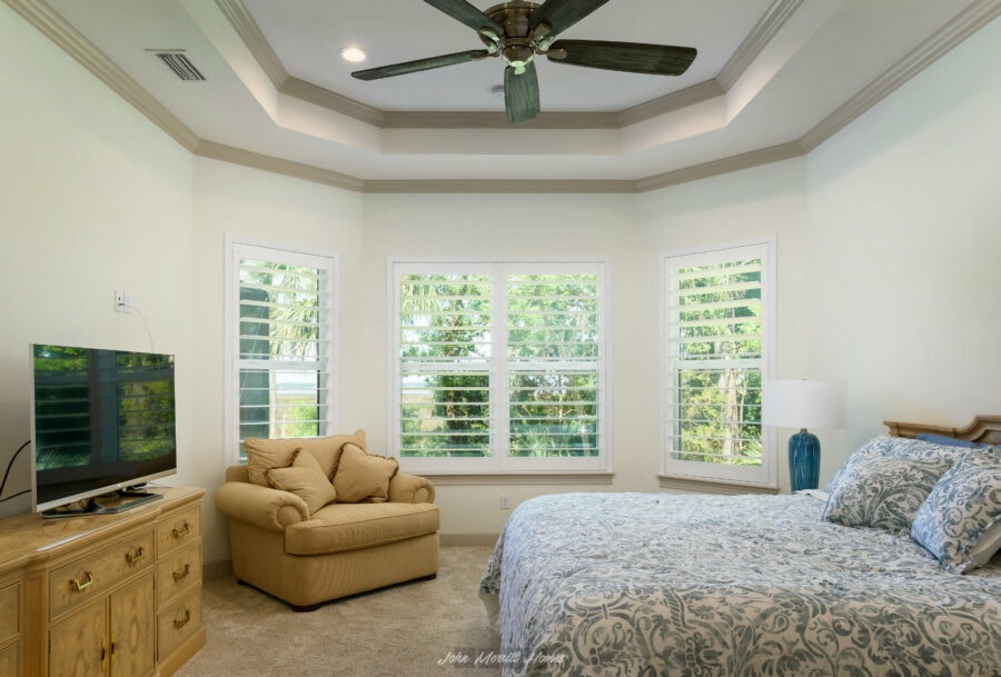 Bright master bedroom featuring large windows with plantation shutters, a beige upholstered armchair, a wooden dresser with a flat-screen TV, and a cozy bed with blue and white geometric bedding.