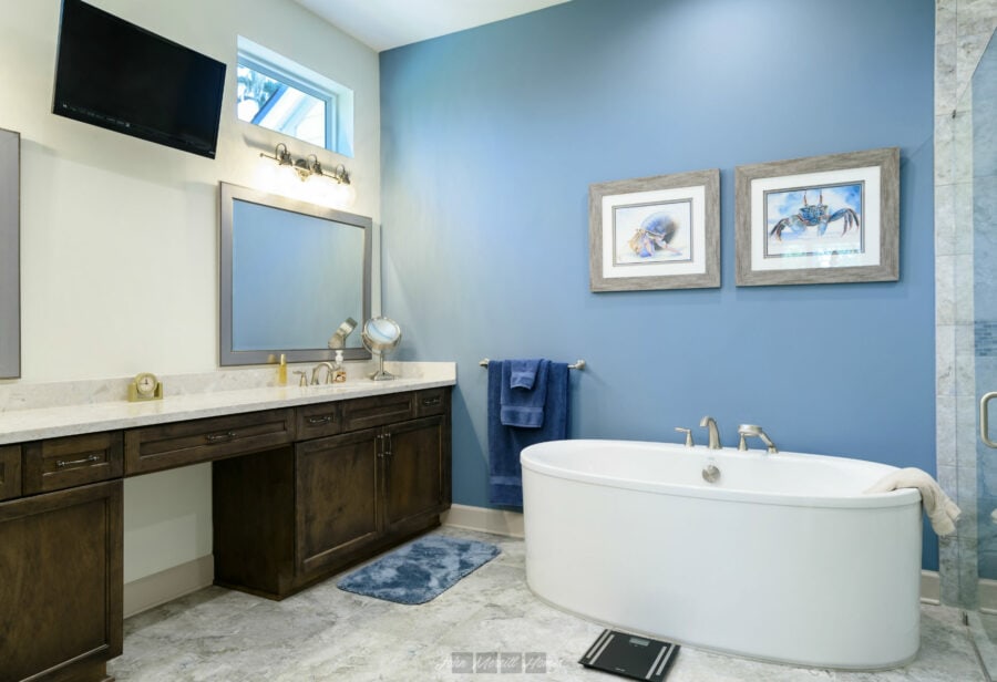 Modern bathroom featuring a large bathtub, dark wood vanity with a marble countertop, wall-mounted mirrors, and framed seaside-themed artwork on a blue accent wall.
