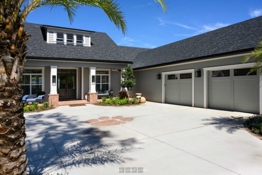 Modern craftsman-style home with a spacious driveway, gray exterior, and a well-maintained front porch featuring lush landscaping and seating areas, under a clear blue sky.