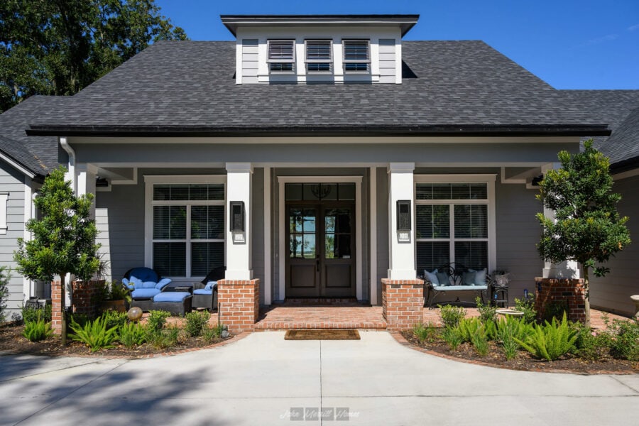 Modern Craftsman style home with gray siding, white trim, and a welcoming front porch featuring brick accents, lush landscaping, and cozy outdoor seating under a bright blue sky.