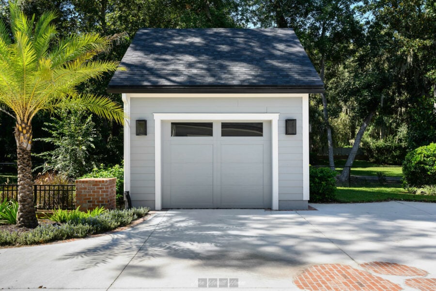 Modern white detached garage with black roof, surrounded by lush greenery and palm trees, featuring a clean concrete driveway and outdoor lighting, exemplifying stylish and functional home design.