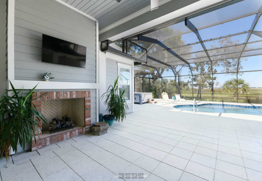 Bright screened-in outdoor pool enclosure with a hot tub, fireplace, and outdoor TV, surrounded by lush greenery and an open landscape, perfect for relaxation and outdoor living.