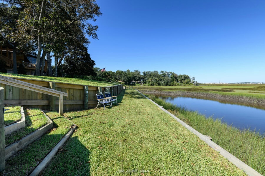 Tranquil waterfront backyard with lush green grass, wooden deck, and scenic view of marshland and waterway under a bright blue sky.