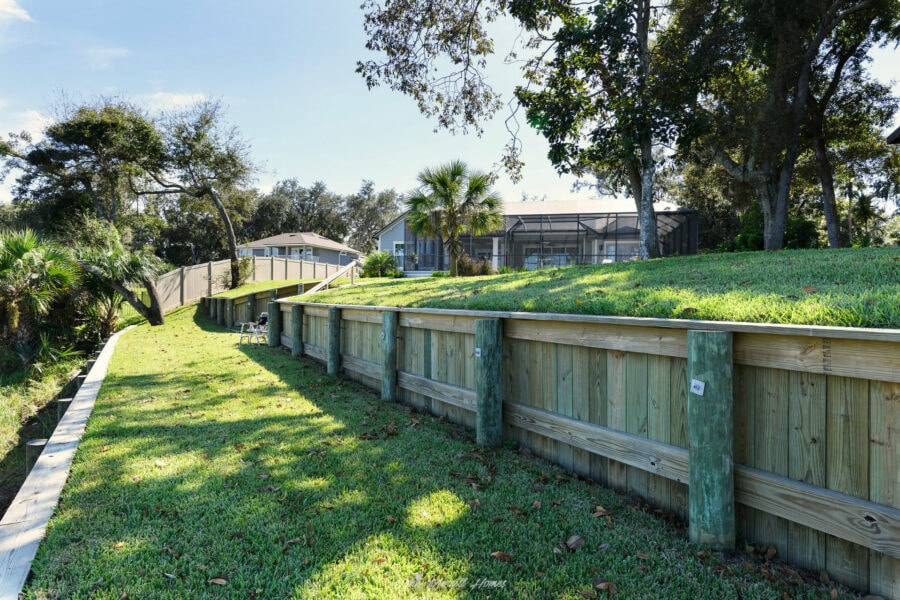 Lush green backyard landscape with a wooden retaining wall, mature trees, and a screened-in porch, showcasing a beautiful outdoor living space at a John Merrill Home.