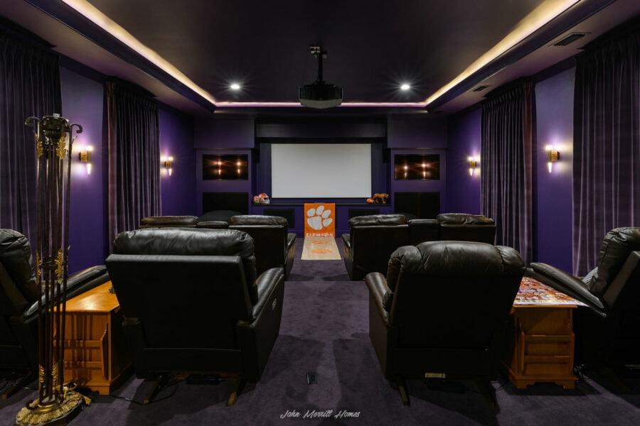 Home theater room with purple walls, black leather recliners, and a large projection screen, featuring ambient lighting and decorative football-themed accents, perfect for an entertainment space.