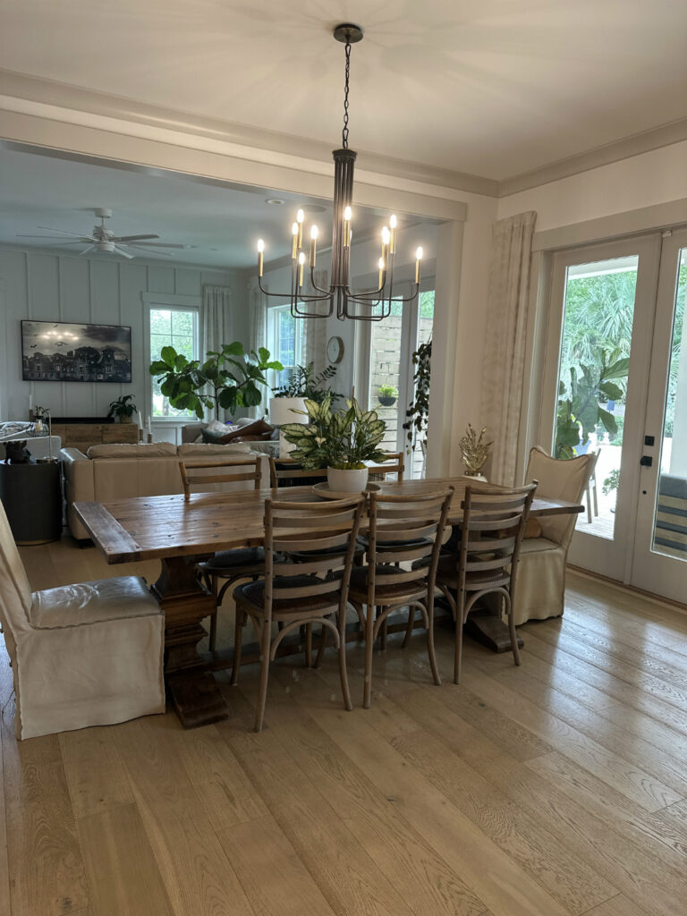 1. A cozy modern dining room with wooden table, eight chairs, large plants, and a black chandelier, featuring large windows and sliding glass door for natural light.