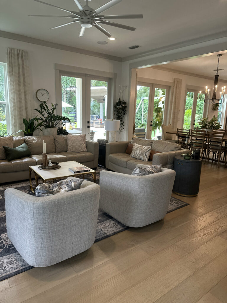 Cozy modern living room with beige sofas, armchairs, large windows, and greenery outside, featuring a ceiling fan, decorative pillows, and a stylish coffee table for family gatherings.