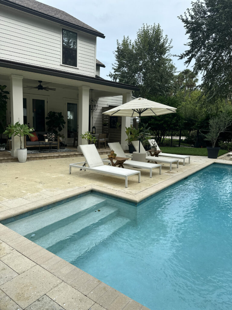 1. Modern backyard with in-ground swimming pool, white lounge chairs, large patio umbrella, and lush greenery surrounding a stylish two-story home.