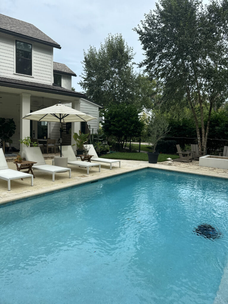 A luxurious backyard with a swimming pool, white lounge chairs, an umbrella, and lush greenery, showcasing modern outdoor living space designed by John Merrill Homes.