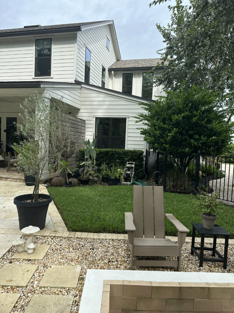 Bright white modern two-story house with large windows, surrounded by lush greenery, a well-maintained lawn, and stylish outdoor seating in a cozy backyard oasis.