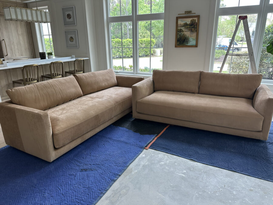 Spacious living room featuring two beige sofas, large windows with greenery outside, and artwork on the walls, creating a bright and inviting atmosphere.