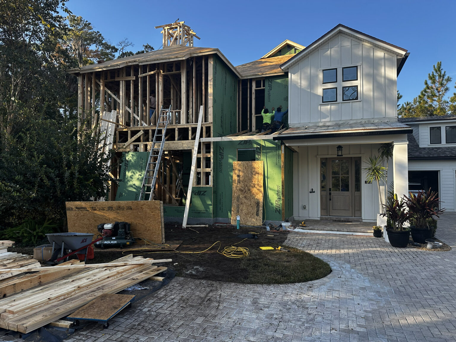 New house under construction with a partly built second story, wooden framework, and workers installing exterior siding; driveway, plants, and construction tools visible.