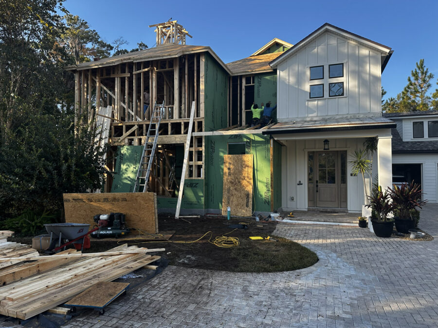 New house under construction with a partly built second story, wooden framework, and workers installing exterior siding; driveway, plants, and construction tools visible.