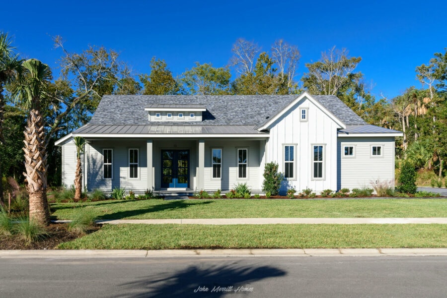 Modern white farmhouse-style home with a front porch, surrounded by lush landscaping and tall trees under a clear blue sky.