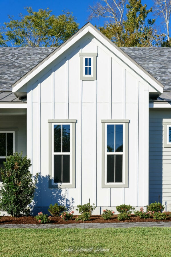 Modern white house with vertical siding, gable roof, and tall windows under a clear blue sky. Well-maintained landscaping highlights the home's charming exterior.