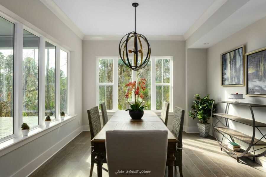 Modern dining room with large windows, a wooden table, and a contemporary black pendant light fixture, creating a bright and welcoming atmosphere; decorated with artwork, plants, and a centerpiece.