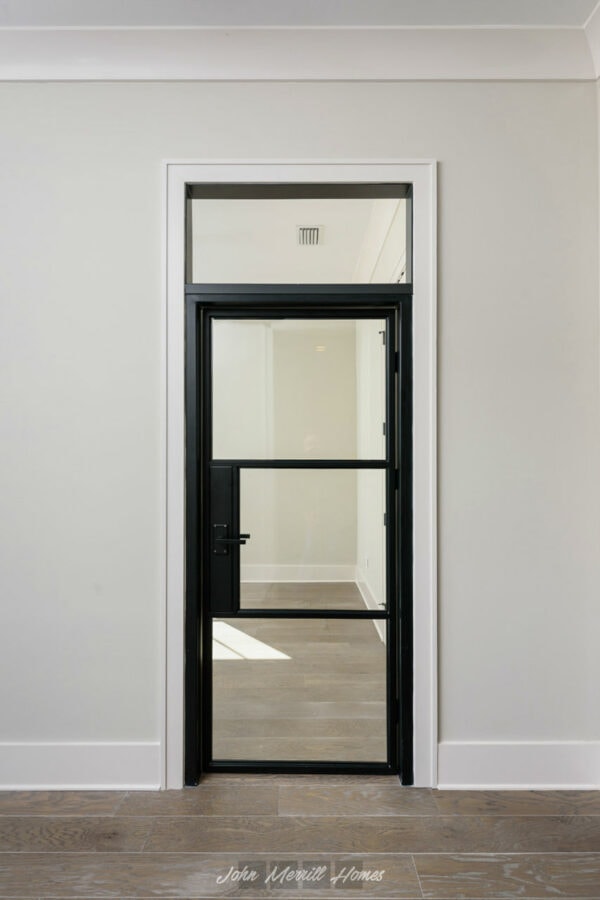 Metal framed glass door with black finish installed in a modern home interior, leading to an adjacent room with natural light and wood flooring.