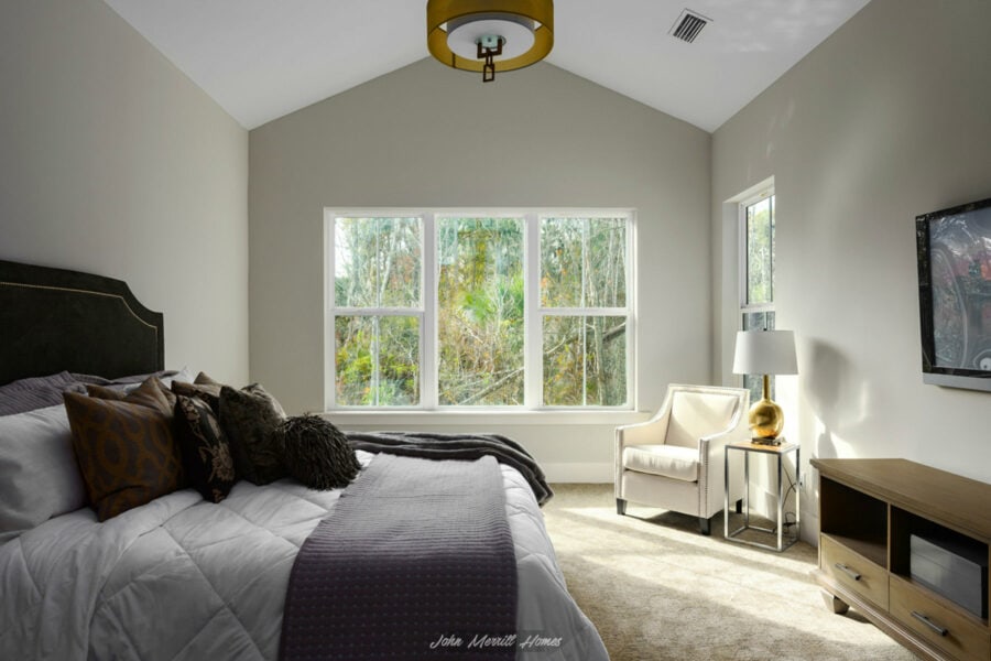 Elegant bedroom with large windows overlooking a lush forest view, featuring a cozy bed with decorative pillows, a modern armchair, stylish side table with gold lamp, and a wall-mounted TV.