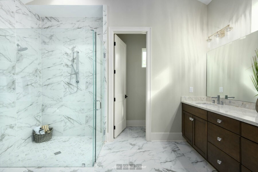 Shower with marble tiles and glass enclosure in a modern bathroom featuring a wooden vanity with marble countertop and a large mirror. Natural light illuminates the space, highlighting elegant design elements.