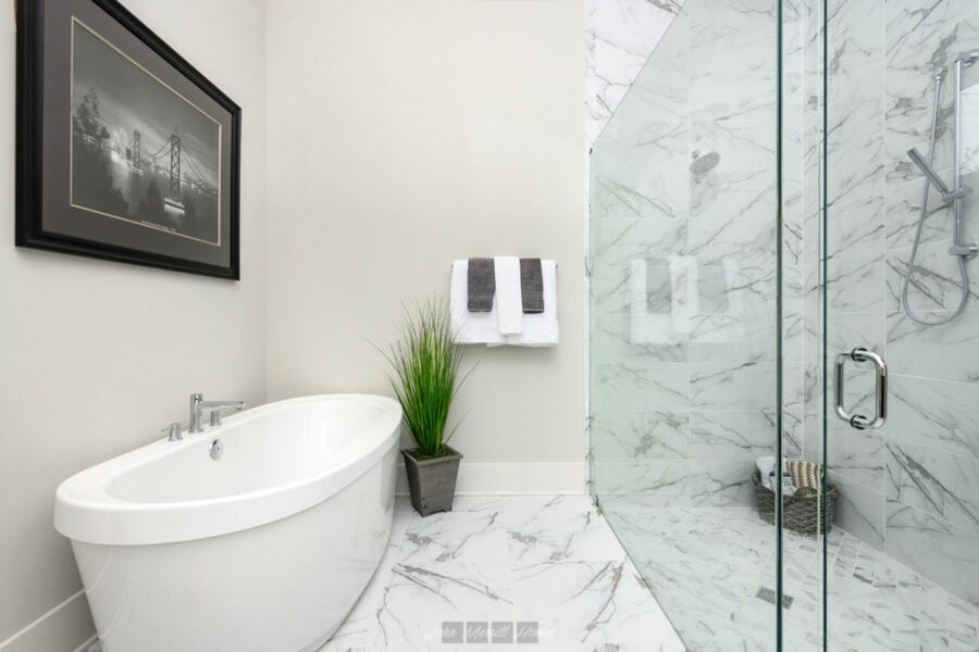 Luxurious modern bathroom featuring a sleek white soaking tub, a marble-tiled shower with glass enclosure, and elegant decor including a framed artwork, a potted plant, and neatly hung towels.