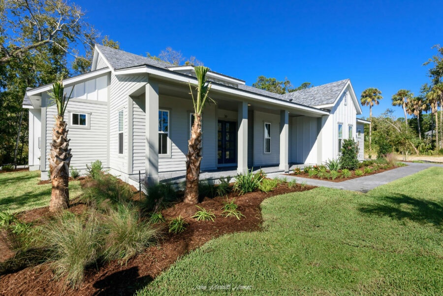 Modern white farmhouse-style home with a spacious front porch, lush landscaped yard with palm trees, and vibrant green grass under a clear blue sky.