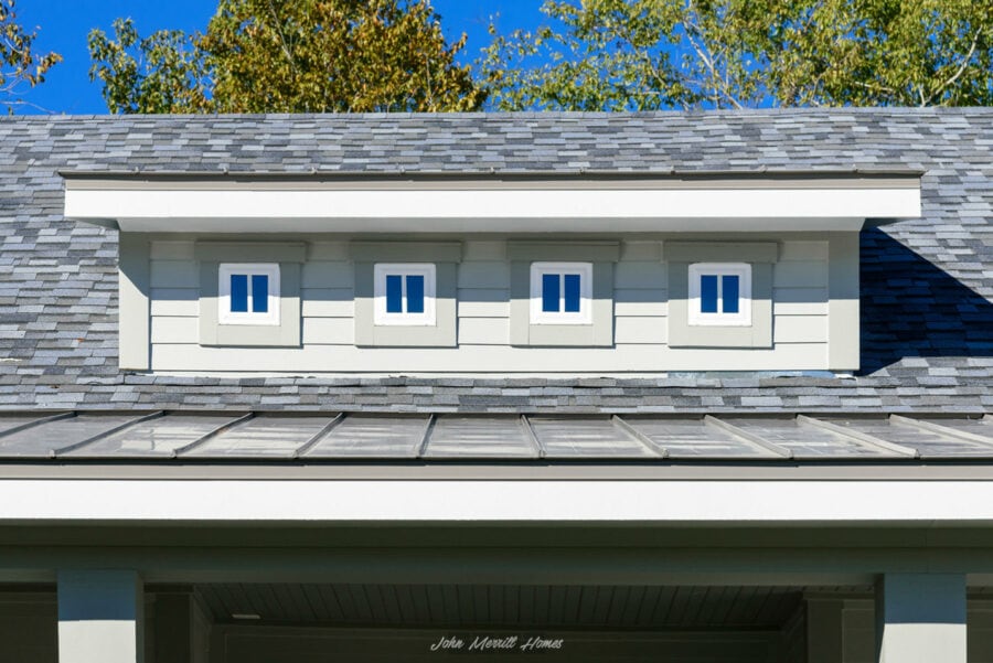 Slim dormer with four small, vertically-paneled windows on a modern home exterior, featuring gray shingle roofing and white siding under a bright blue sky.