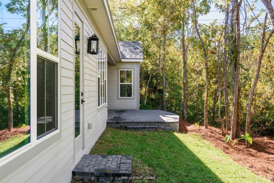 Modern home exterior with a cozy backyard patio, surrounded by lush trees and greenery, featuring a sliding glass door, white siding, and black outdoor lighting fixtures.