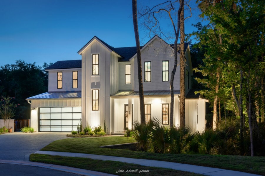 Luxurious modern white home illuminated at dusk with large windows, surrounded by lush greenery, showcasing contemporary architecture and elegant outdoor lighting.