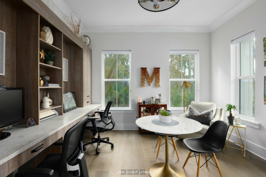 Modern home office with built-in wooden desk and shelves, black swivel chairs, a white round table with gold base, comfortable armchair, and large windows with greenery views.