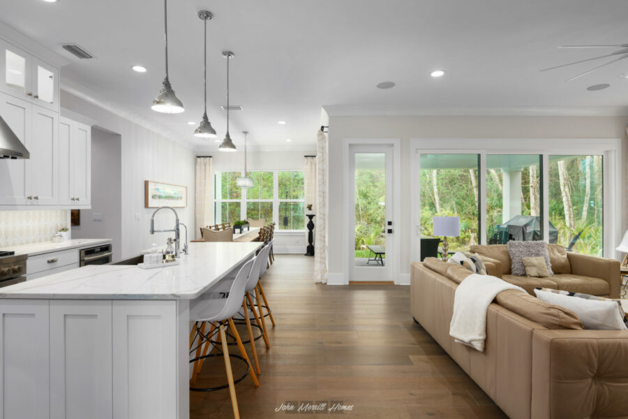 Bright open-concept kitchen and living area with white cabinetry, marble countertops, wooden flooring, and large windows overlooking a lush backyard.