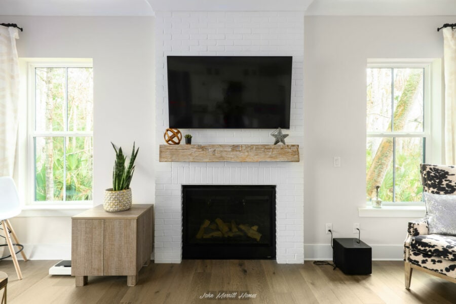 Modern living room with white brick fireplace, mounted flat-screen TV, large windows with natural light, light wood flooring, and contemporary decor from John Merrill Homes.