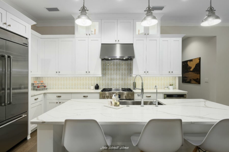 Bright white modern kitchen with a large central island, stainless steel appliances, pendant lighting, and a backsplash with a geometric pattern, designed by John Merrill Homes.