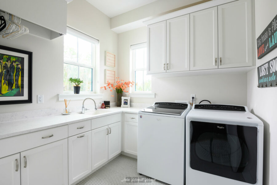 Laundry room with white cabinets, a washing machine, a dryer, a sink, and decorative plants, sunlight streaming through windows, creating a bright and clean space.