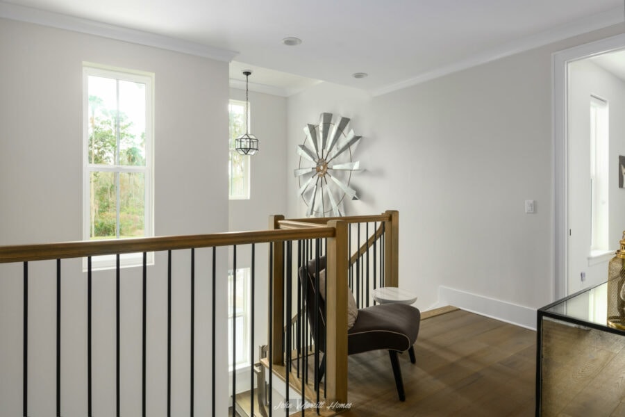 Modern staircase landing with white walls, large windows, decorative windmill wall art, wooden floor, and contemporary furniture in a stylish home interior.