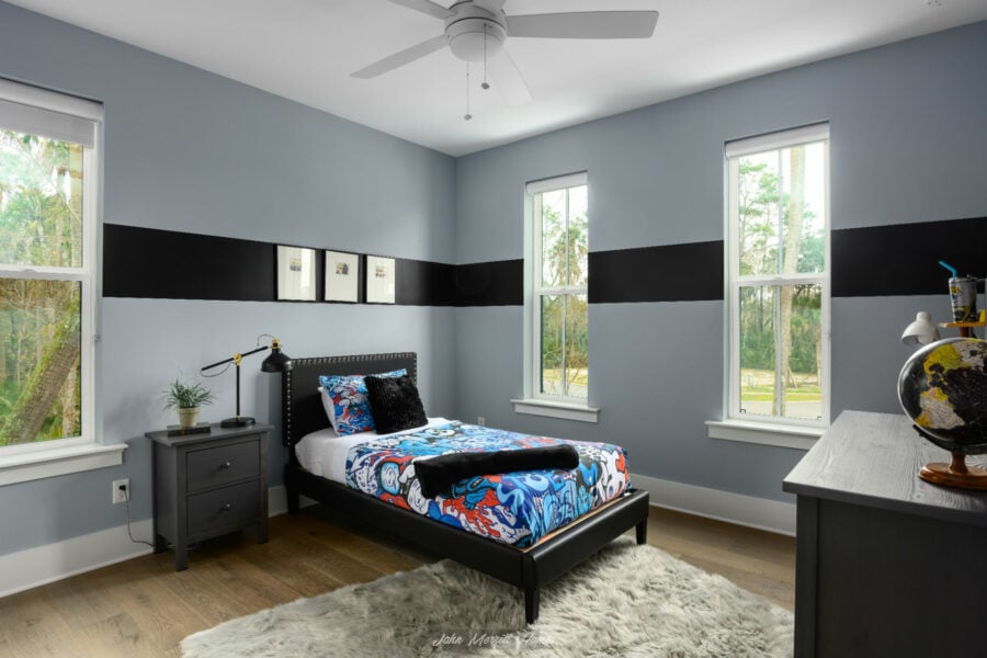 Modern bedroom with light blue walls accented by a bold black stripe, featuring a black bed frame with colorful bedding, a gray nightstand with a lamp, and large windows letting in natural light.