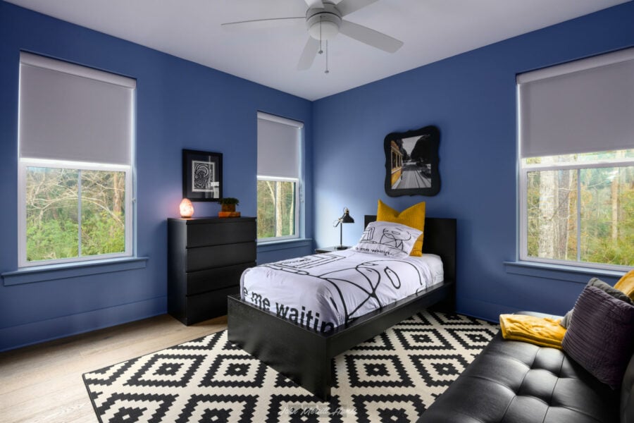 Bright blue bedroom with large windows, black furniture, and modern decor, featuring a stylish bed, colorful pillows, and a geometric rug, creating a cozy and vibrant atmosphere.